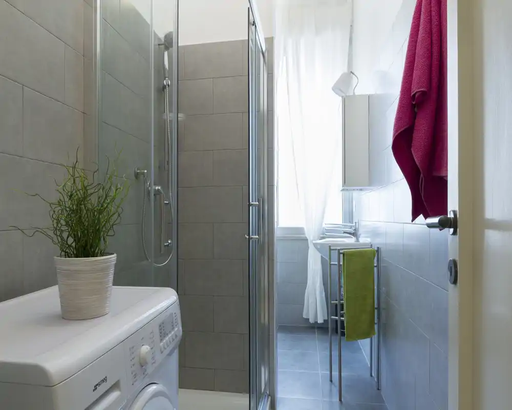 Bagno moderno e luminoso con piastrelle, box doccia in vetro, lavatrice e lavabo compatto — spazio pulito e ben illuminato, ideale per affitti.