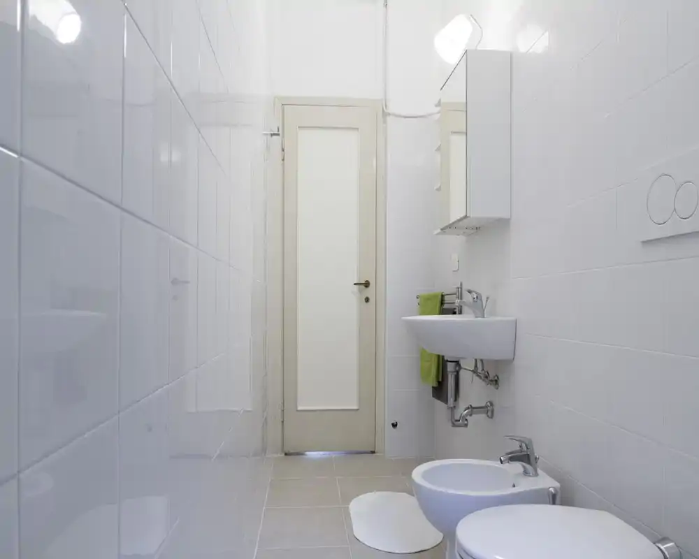 Bagno pulito e stretto con piastrelle bianche, lavabo sospeso, bidet e WC, e una porta satinata sul fondo. Stile luminoso e minimal ideale per annunci.