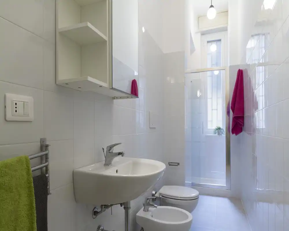 Bagno moderno e luminoso con lavabo a parete, bidet e WC, porta doccia in vetro e piastrelle bianche alle pareti.