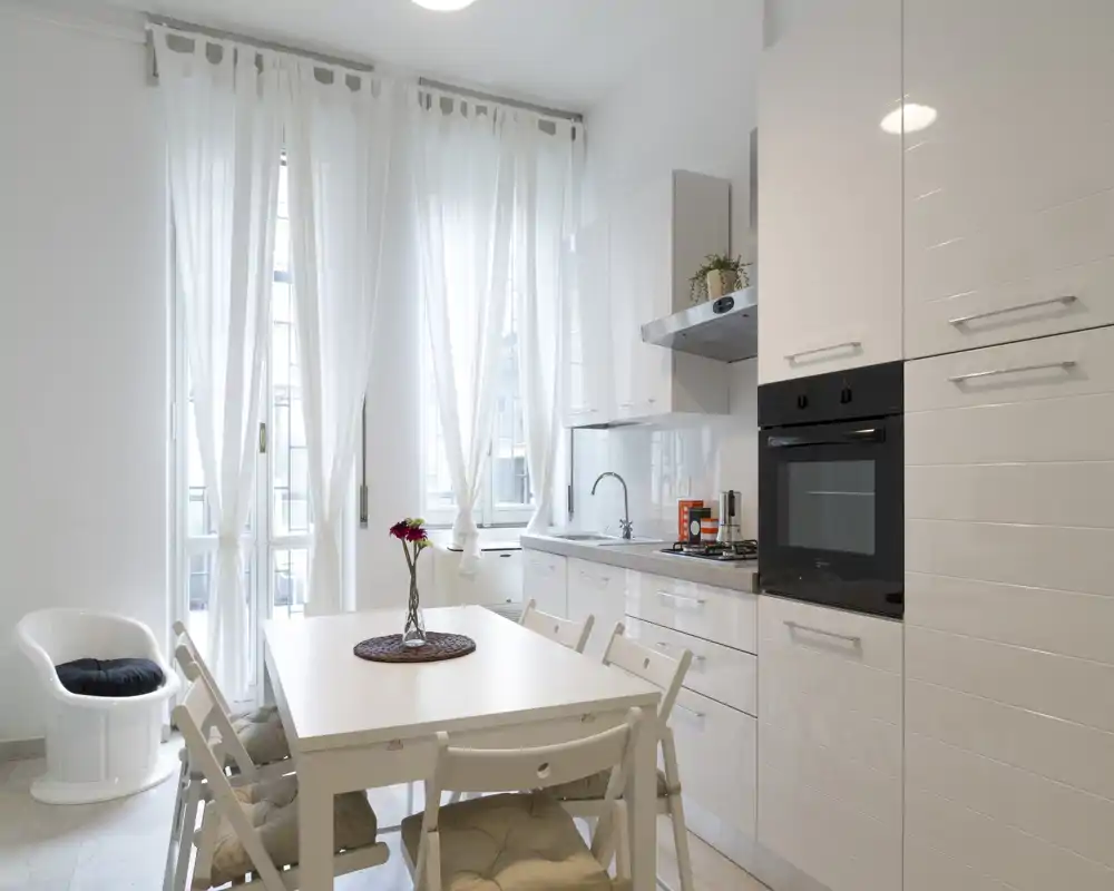 Cucina moderna e luminosa con elettrodomestici integrati, tavolo e sedie bianchi e finestre alte con tende chiare che diffondono luce naturale.