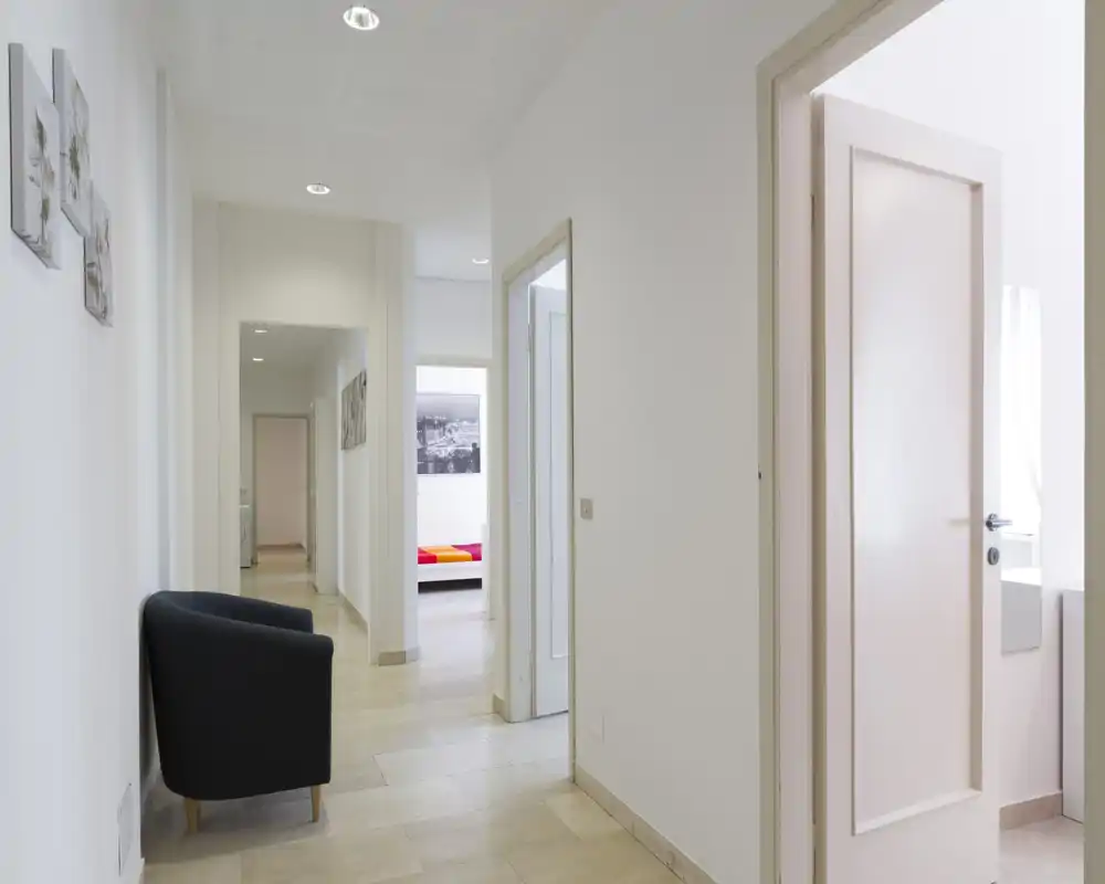 Ingresso luminoso e minimal con pavimento piastrellato, una poltroncina e più porte che conducono ad altre stanze — pulito e ben illuminato.