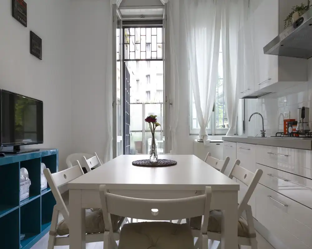 Zona pranzo luminosa e moderna integrata con angolo cottura: tavolo bianco per sei persone, vaso come centrotavola e molta luce naturale da ampie finestre e una porta sul balcone.
