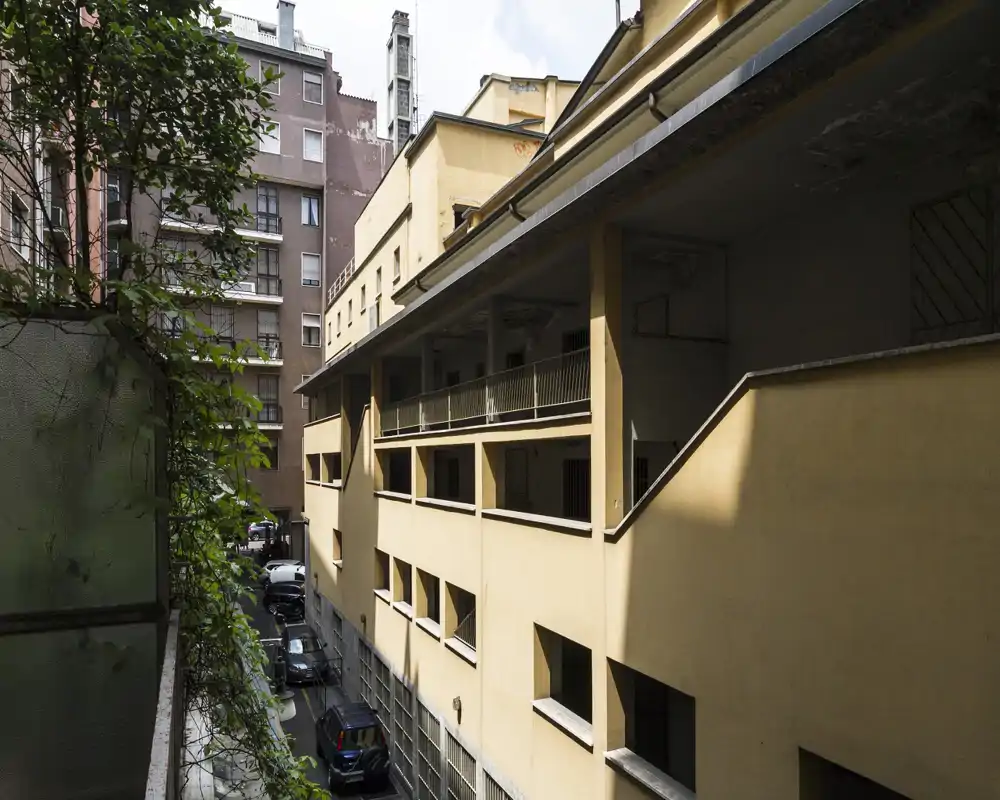 Vista su un cortile interno e palazzi adiacenti da un balcone: facciate gialle, corridoi esterni e auto parcheggiate nel vicolo sottostante.