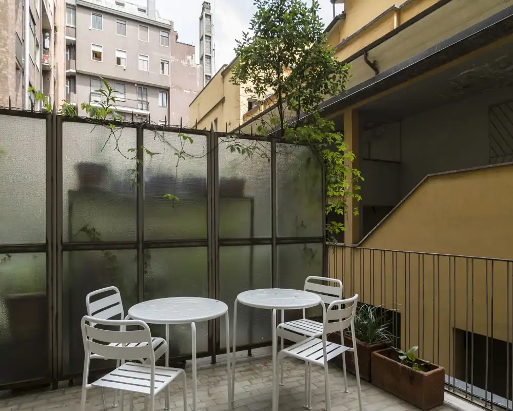 Piccolo balcone privato con due tavolini rotondi, quattro sedie e vasi con piante; riparato da pannelli in vetro satinato e affacciato sul cortile interno — perfetto per il caffè mattutino.