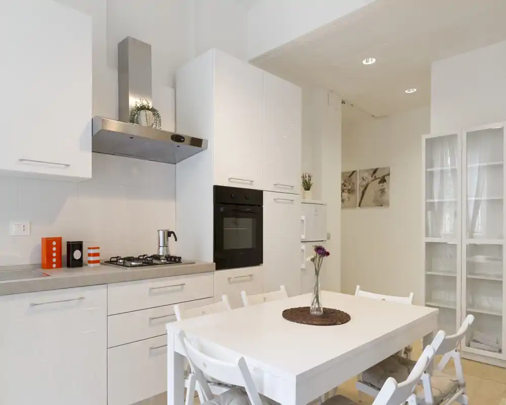 Cucina moderna e luminosa con forno incassato, piano cottura a gas e tavolo da pranzo bianco — stile minimal pulito e un vaso come centrotavola che crea atmosfera.