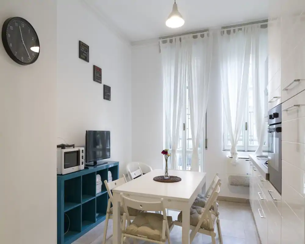 Area pranzo luminosa e minimale con tavolo bianco e sedie pieghevoli imbottite, adiacente a una cucina compatta e grandi finestre con tende leggere.