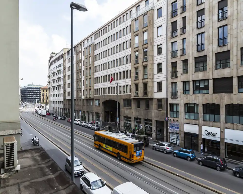 Veduta urbana dalla finestra di un edificio: strada a più corsie con binari del tram, veicoli incluso un autobus giallo e palazzi commerciali adiacenti.