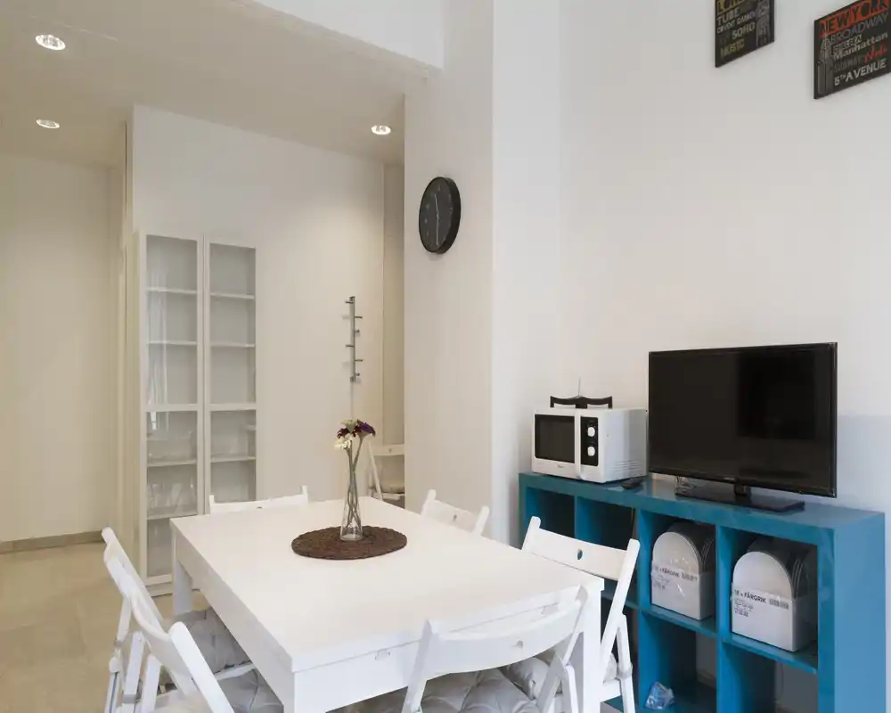 Zona pranzo luminosa e moderna con tavolo bianco e sedie pieghevoli, un piccolo centro tavola e un mobile contenitore con elettrodomestici e TV.