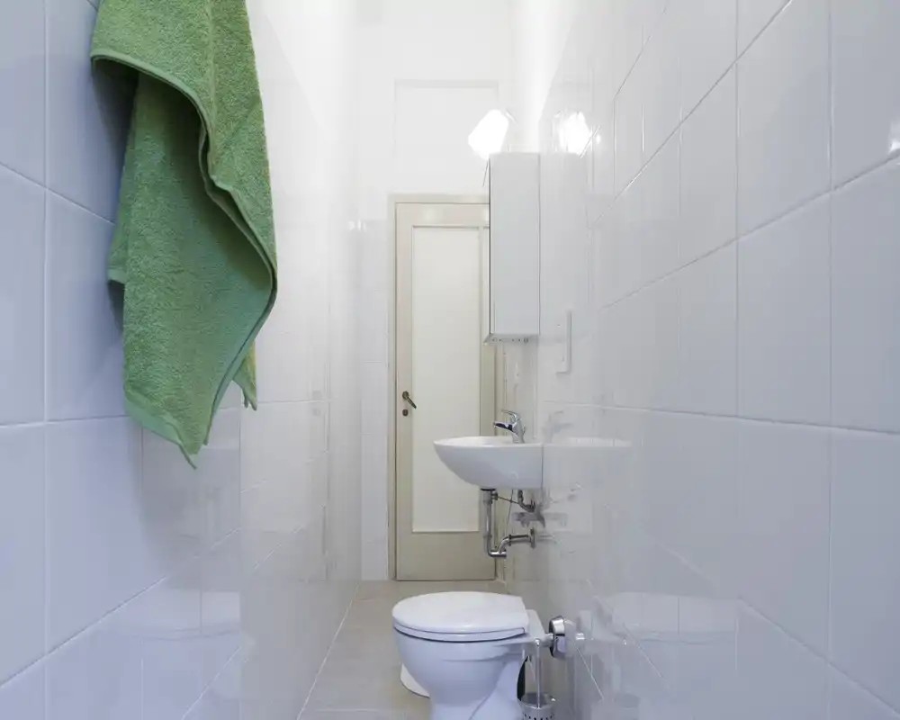 Bagno stretto e luminoso con piastrelle bianche, lavabo a parete, WC e un asciugamano verde appeso — ambiente pulito e compatto, ideale per affitti brevi.