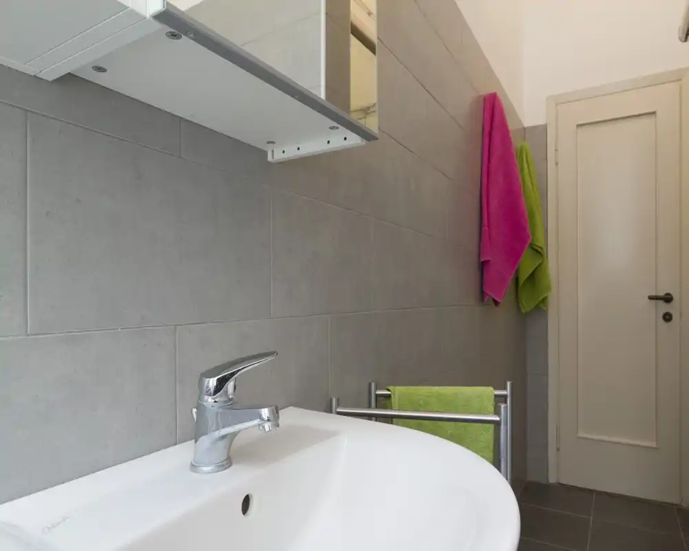 Bagno moderno e compatto con lavabo bianco, rubinetto cromato e pensile con specchio; asciugamani colorati che danno un tocco vivace.