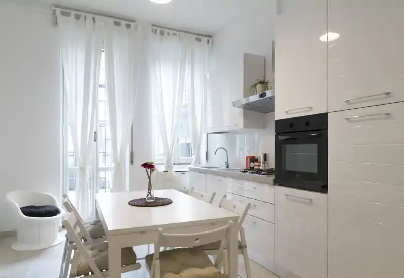 Cucina moderna e luminosa con elettrodomestici integrati, tavolo e sedie bianchi e finestre alte con tende chiare che diffondono luce naturale.