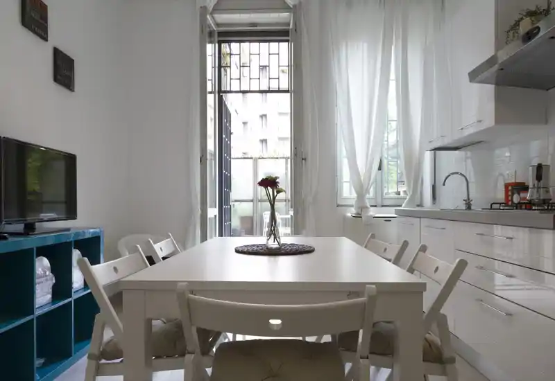 Zona pranzo luminosa e moderna integrata con angolo cottura: tavolo bianco per sei persone, vaso come centrotavola e molta luce naturale da ampie finestre e una porta sul balcone.