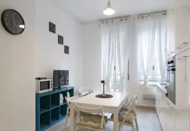 Area pranzo luminosa e minimale con tavolo bianco e sedie pieghevoli imbottite, adiacente a una cucina compatta e grandi finestre con tende leggere.