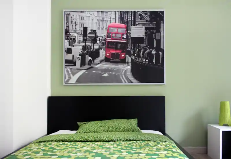 Dormitorio moderno y luminoso con una cama doble visible, ropa de cama verde con estampado y un gran cuadro enmarcado sobre la cabecera.