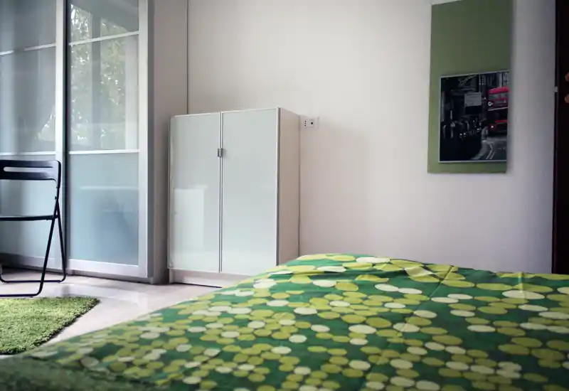 Dormitorio moderno y luminoso con edredón verde estampado, armario con puertas esmeriladas y luz natural de una ventana grande; estilo sencillo y fresco, ideal para anuncios.