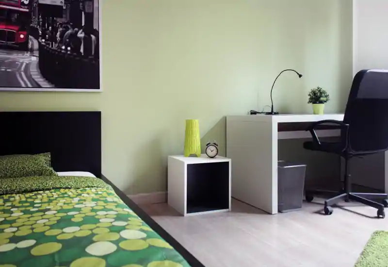 Dormitorio moderno y luminoso con colcha verde estampada, mesita cúbica y escritorio blanco con silla de trabajo — limpio, minimalista y bien iluminado.