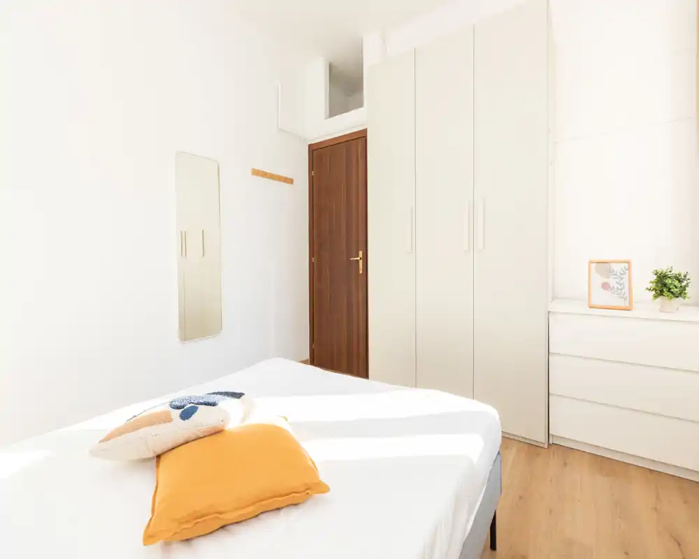 Chambre lumineuse et minimaliste avec lit bien fait, sol en bois chaleureux et placard intégré. La lumière naturelle et les coussins décoratifs apportent une ambiance accueillante.