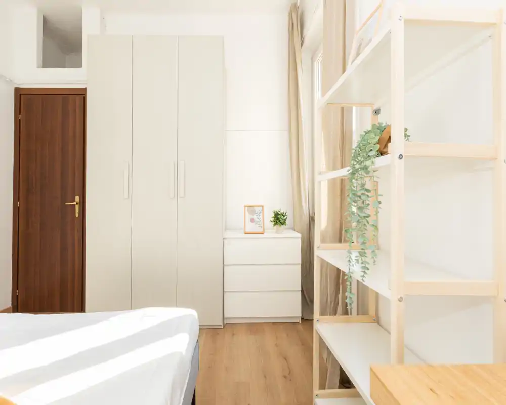 Chambre minimaliste et lumineuse avec un lit au premier plan, une armoire blanche et une commode, et un sol en bois clair. La lumière naturelle et les textiles neutres donnent une atmosphère calme et aérée.