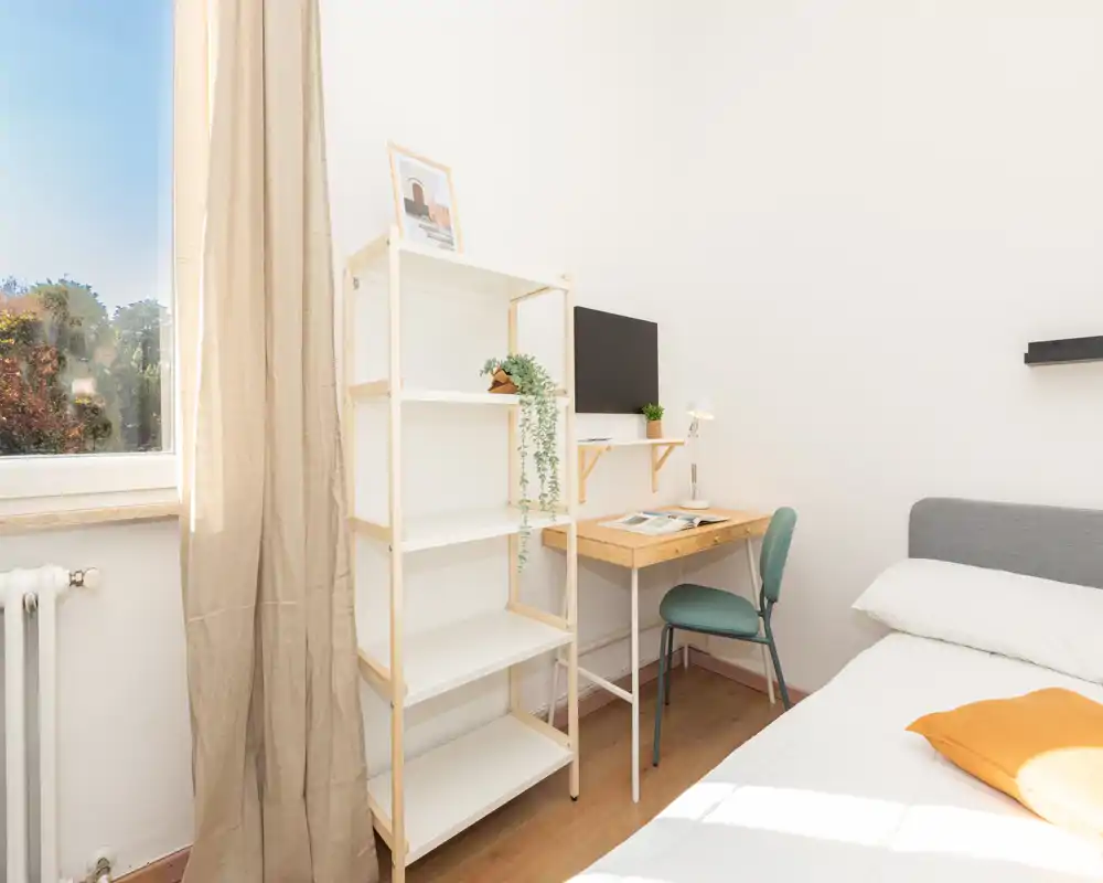 Chambre lumineuse et minimaliste avec un lit confortable, un petit bureau en bois et des étagères près d'une grande fenêtre qui apporte beaucoup de lumière naturelle.