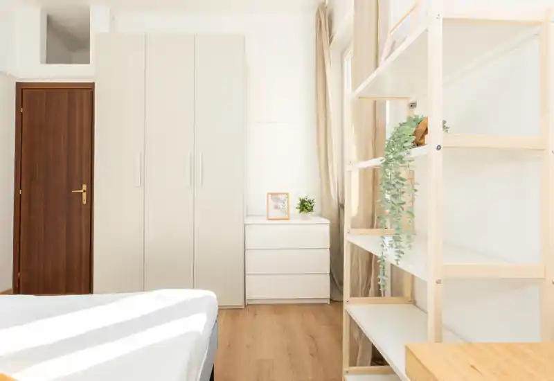 Camera da letto luminosa e minimal con il letto in primo piano, un armadio bianco e un comò, pavimento in legno chiaro. La luce naturale e i tessuti neutri creano un ambiente sereno e arioso.