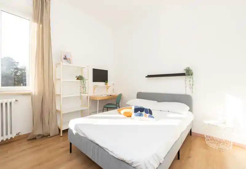 Camera da letto luminosa e minimale con letto matrimoniale, cuscini decorativi, una piccola scrivania e scaffalatura vicino alla finestra che lascia entrare molta luce naturale.