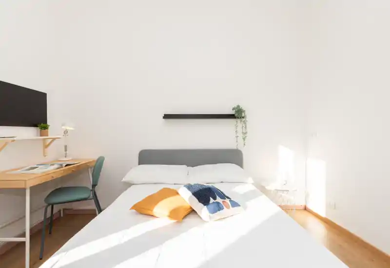 Camera da letto luminosa e minimalista con letto matrimoniale al centro, biancheria bianca, cuscini colorati e una piccola scrivania a muro — perfetta per l'annuncio di un alloggio.