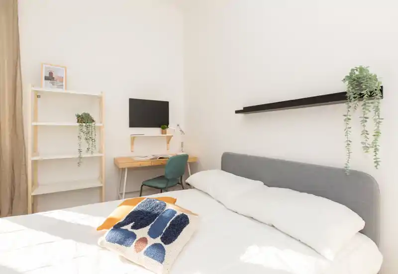 Camera da letto luminosa e minimale con letto matrimoniale ben rifatto, cuscino decorativo e piante sospese; piccolo angolo studio con scrivania e scaffale.