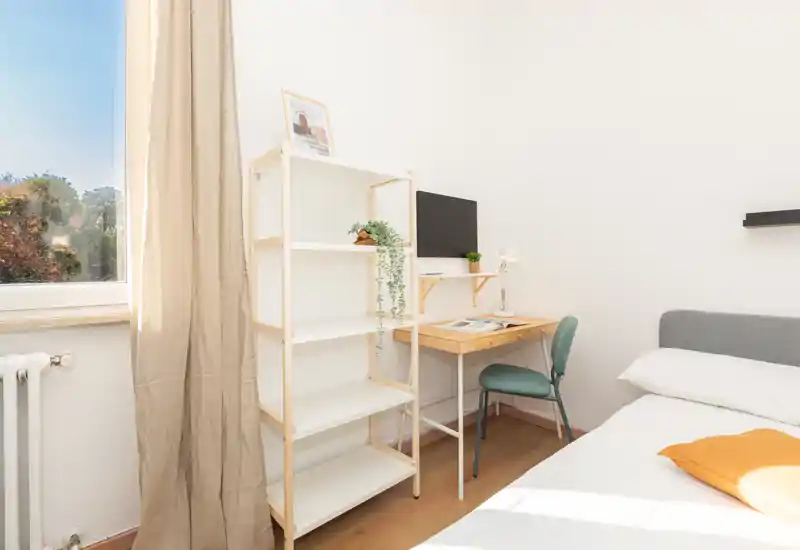 Camera da letto luminosa e minimal con un comodo letto, una scrivania in legno compatta e scaffalatura accanto a una grande finestra che inonda la stanza di luce naturale.