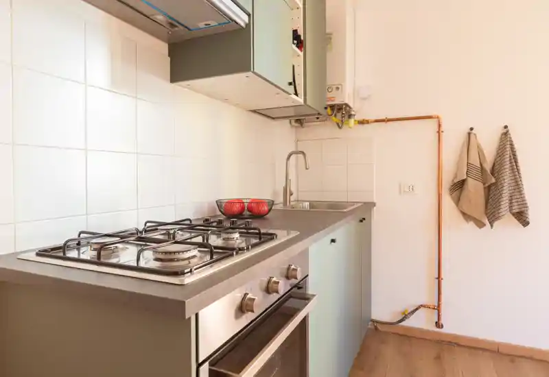 Cucina moderna compatta con piano cottura a gas, forno incassato, lavello in acciaio e pavimento in legno caldo — arredata con una ciotola di frutta e asciugamani appesi.