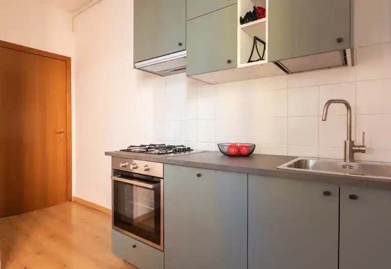 Cucina moderna compatta con piano cottura a gas e forno integrato, lavello in acciaio e mobili verdi pastello. Piano di lavoro pulito con una ciotola di frutta decorativa e pavimento in legno che dona calore.