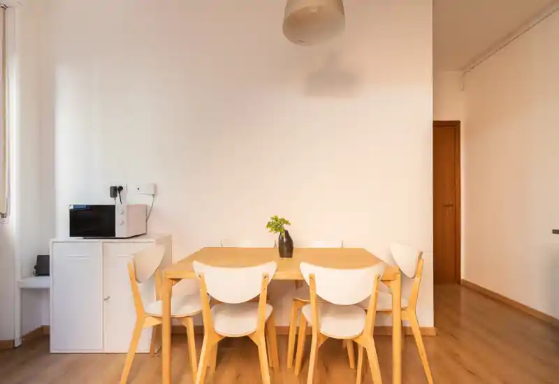 Zona pranzo luminosa e minimale con tavolo in legno, sei sedie abbinate e un piccolo vaso al centro — ideale per annunci di appartamenti compatti.