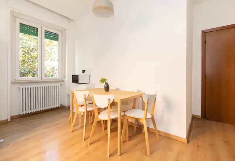 Zona pranzo luminosa e moderna con tavolo in legno e sei sedie vicino a una grande finestra, arredamento minimal e pavimento in legno caldo.