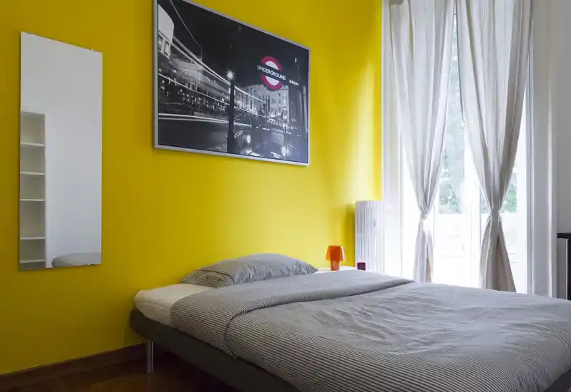 Camera da letto moderna e luminosa con letto matrimoniale contro una parete gialla vivace, ampia finestra con tende leggere, lampada da comodino e arredo minimalista.