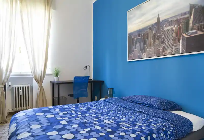 Bright, modern bedroom with a double bed dressed in blue patterned bedding, a bedside lamp and a desk by a window letting in natural light.