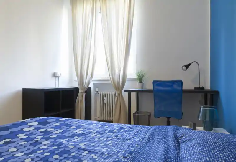 Bright, simply styled bedroom with patterned blue bedding, a study desk and natural light from a window — comfortable and functional for short stays.