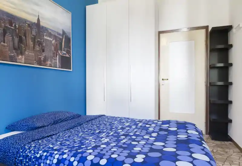 Bright, tidy bedroom with a double bed dressed in patterned blue linens, a white wardrobe and striking blue accent wall — a compact, modern space ideal for couples or solo travelers.