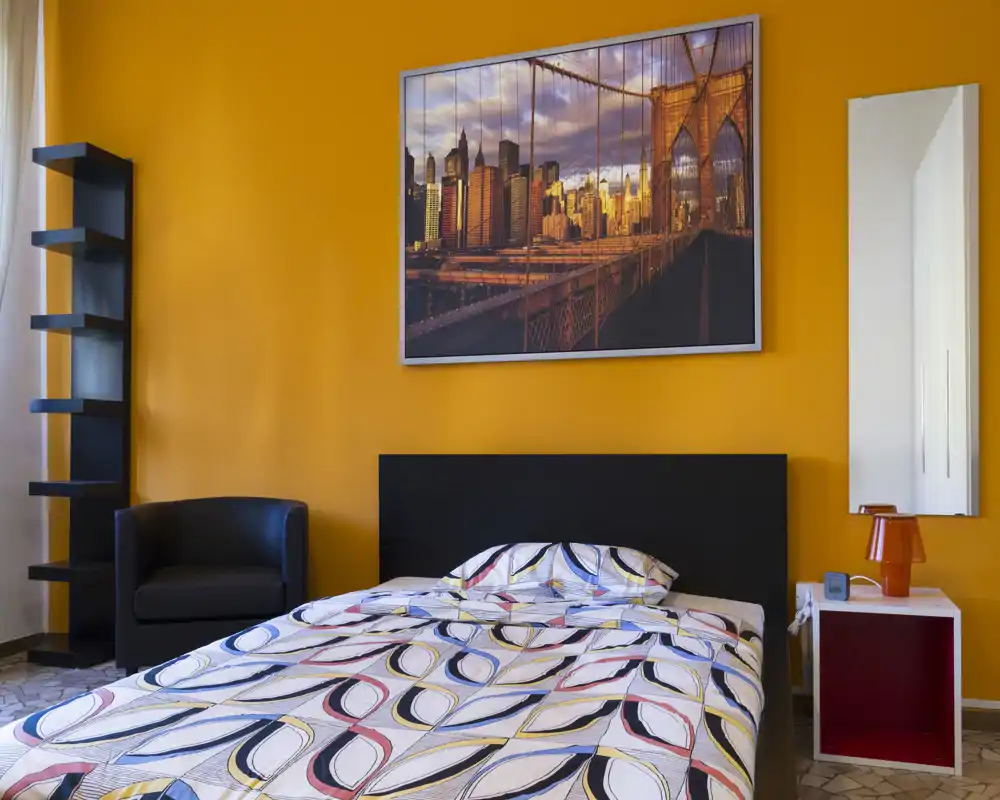 Bright, stylish bedroom with a patterned double bed, bold orange accent wall, framed city artwork, armchair and a bedside cabinet with lamp — modern and inviting.