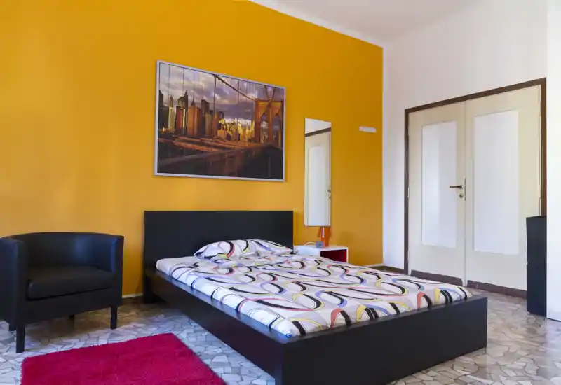Bright, modern bedroom with a low black platform bed against a warm orange accent wall, patterned bedding, a black armchair and a red rug — a cozy, well-lit space ideal for short-term rentals.
