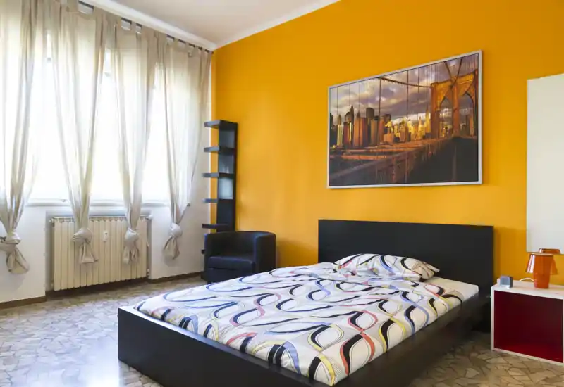 Sunny bedroom with a double bed against a bold orange accent wall, large windows with sheer curtains, and modern decorative touches including a framed city print and a small armchair.