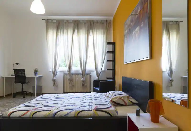Bright, modern bedroom with a double bed facing a large window, warm orange accent wall and a tidy workspace in the corner.