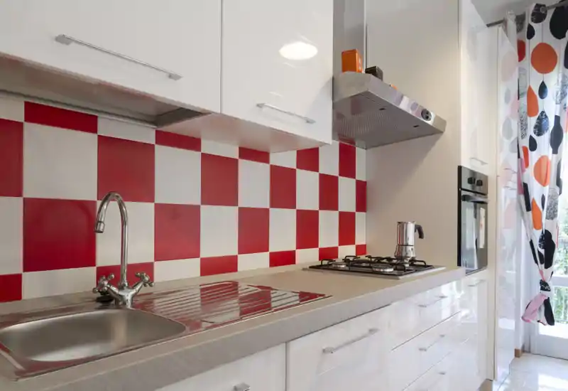Bright modern kitchen with a red-and-white checkered backsplash, stainless sink, gas hob and built-in oven; cheerful curtains let natural light in.