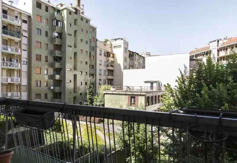 Sunny private balcony overlooking a courtyard with planters on the railing and surrounding apartment buildings; pleasant urban view and good natural light.
