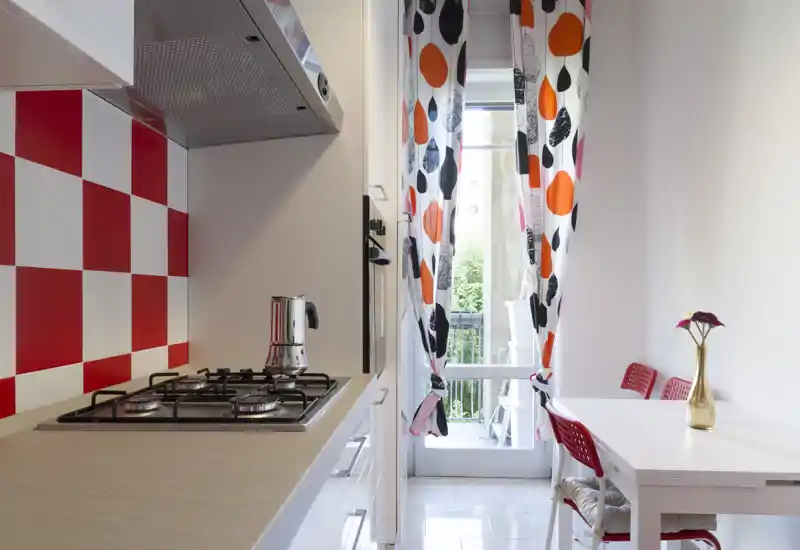 Bright compact kitchen with a four-burner gas hob, red-and-white tiled backsplash and a small dining table by a door to a balcony; colorful curtains and a decorative vase add personality.