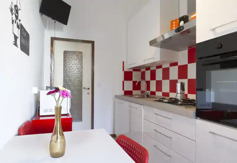 Compact modern kitchen with white cabinets and distinctive red-and-white checker backsplash, gas hob and built-in oven; small dining table with red chairs and a decorative vase.