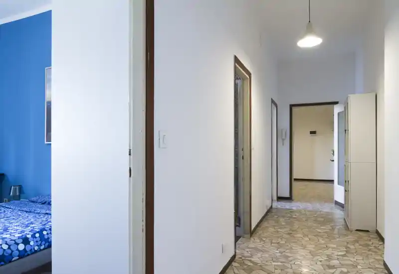 Long entrance corridor with tiled floor, white walls and a visible bedroom doorway to the left. Clean, well-lit transitional space suitable for listing the apartment layout.