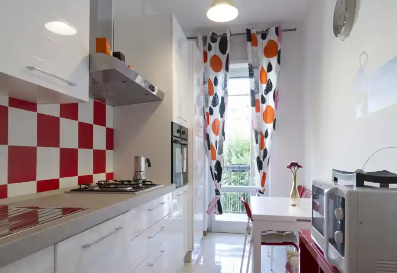 Bright modern kitchen with white high-gloss cabinets, red-and-white checker backsplash, gas hob and a small dining table by a large window with colorful curtains.