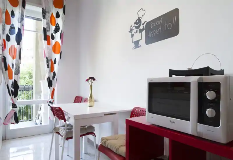 Compact kitchen/dining corner with a white table, red chairs and a microwave on a red shelf; bright natural light and playful patterned curtains create a cheerful feeling.