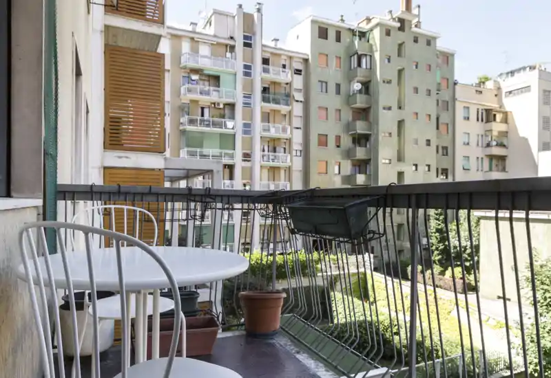 Balcón urbano soleado con una mesa redonda pequeña, sillas y macetas con vistas a edificios vecinos — un rincón acogedor para el café de la mañana.