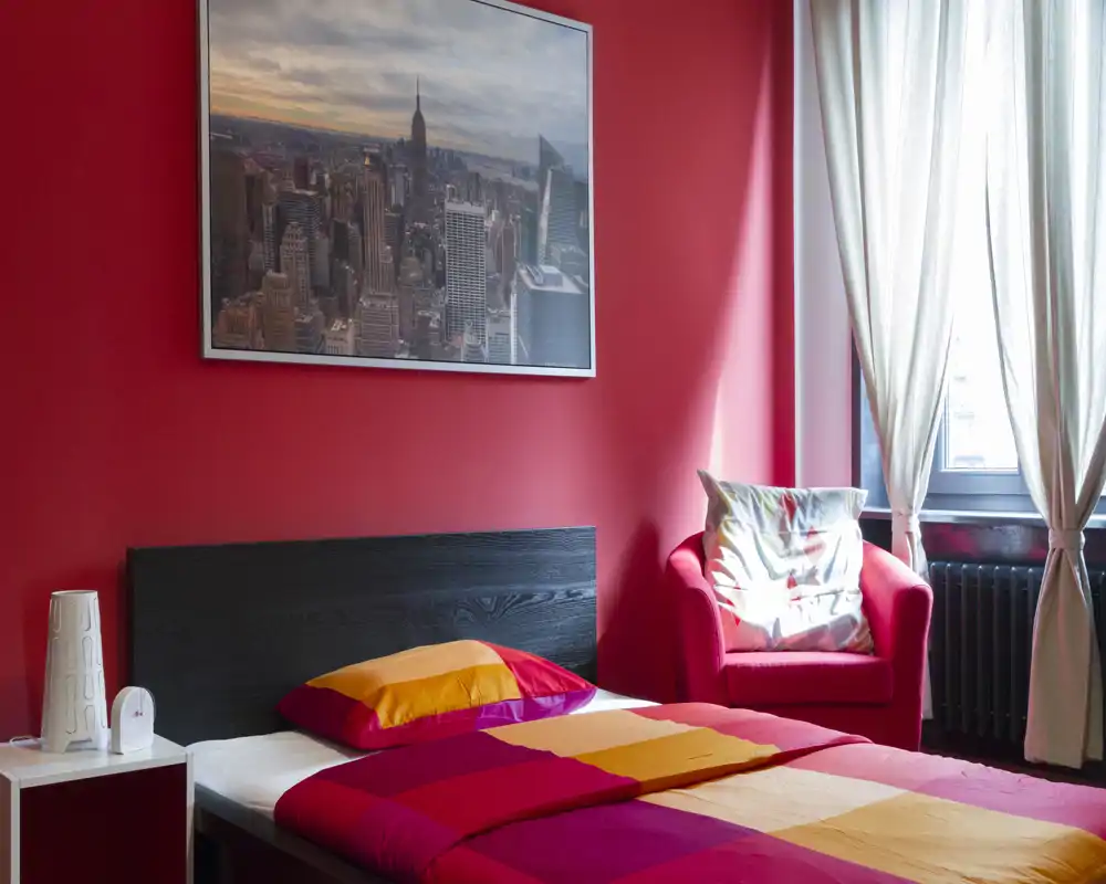 Camera da letto luminosa e colorata con letto matrimoniale contro una parete rossa e una poltrona comoda vicino alla finestra. Biancheria vivace, quadro con città e luce naturale soffusa.