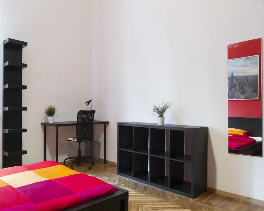 Camera da letto moderna e luminosa con copriletto colorato, scrivania compatta e scaffalature — ideale per studenti o single.